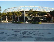 Veduta esterna di un grande stadio moderno con struttura ad anello e travi bianche a traliccio sul tetto, circondato da alberi e facciate vetrate. In primo piano si vede un’ampia piazza pavimentata con decorazioni a mosaico e aiuole basse.