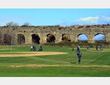 Campo da golf con ampie distese di erba e una buca segnalata da bandierina, con sullo sfondo un lungo acquedotto in pietra con grandi arcate. Terreno leggermente ondulato in ambiente aperto, con vegetazione bassa e cielo azzurro.