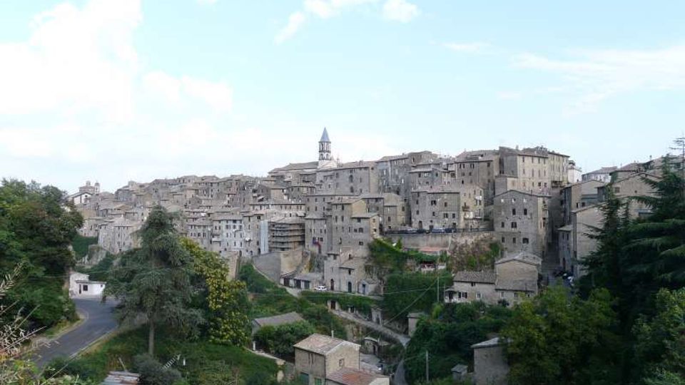 Vista panoramica di un borgo collinare con case in pietra addossate tra loro e un campanile che svetta al centro. In primo piano si vedono vegetazione, piccoli edifici rurali e una strada curvilinea ai margini del paese.