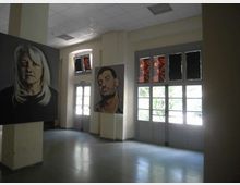 Interno di una galleria o sala espositiva con pavimento lucido e soffitto a pannelli, illuminata da grandi porte-finestre a vetri e finestre alte. Su pilastri e pareti sono appesi grandi quadri con ritratti, disposti lungo lo spazio aperto.