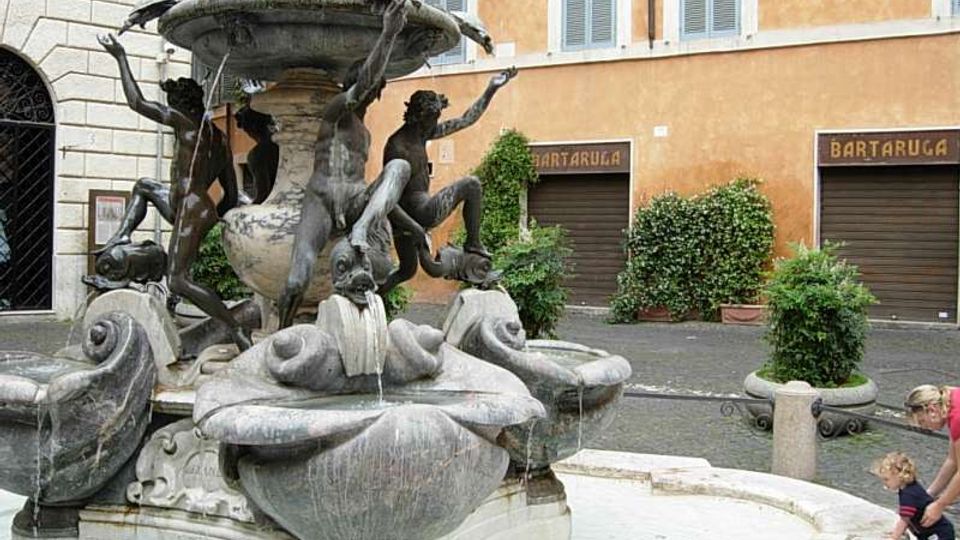 Fontana barocca in una piazza cittadina, con vasca circolare e sculture di figure e delfini che sorreggono una grande conchiglia centrale da cui sgorga l’acqua. Sullo sfondo si vedono edifici storici intonacati con finestre e serrande, e alcune piante in vaso lungo il marciapiede.
