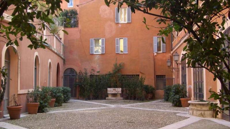 Cortile interno pavimentato in ciottoli con aiuole e vasi di piante lungo i lati, circondato da edifici intonacati color terracotta con finestre e persiane. Sullo sfondo si vede un arco d’ingresso e al centro una panchina in pietra, con rami di alberi che incorniciano la scena.