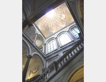 Interno di un edificio storico con grande vano scala in pietra, balaustre e colonne decorate, visto dal basso verso una cupola a cassettoni con lucernario centrale. Alte finestre ad arco illuminano le pareti ornate con stucchi e motivi classici.