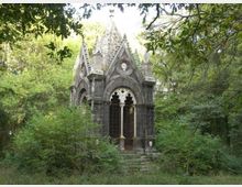 Piccola cappella in pietra in stile gotico, con archi a sesto acuto, finestre traforate e cuspidi decorative, situata in un’area boschiva. Vegetazione fitta e rami degli alberi circondano l’edificio e una breve scalinata conduce all’ingresso.