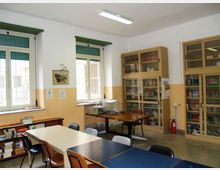 Aula scolastica con tavoli e sedie, pareti bianche con fascia gialla e due grandi finestre con tapparelle verdi. Sul lato destro ci sono armadi vetrati pieni di libri e materiali, con una stampante su un tavolo e un estintore vicino alla parete.