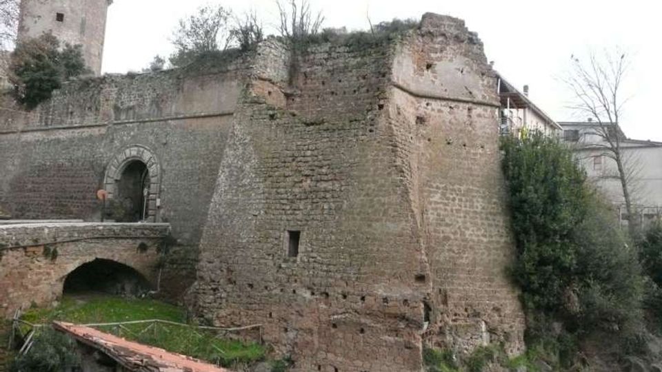 Ruderi di una fortificazione in muratura con grande bastione in pietra e torre cilindrica sullo sfondo. In primo piano si vede un arco di ponte e un tetto in coppi, con vegetazione e pareti rocciose attorno alle mura.