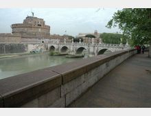 Veduta del Tevere con un ponte in pietra a più arcate e, sullo sfondo, una grande fortezza cilindrica in muratura. In primo piano si vede una lunga banchina con muro in pietra e un viale alberato lungo il fiume.
