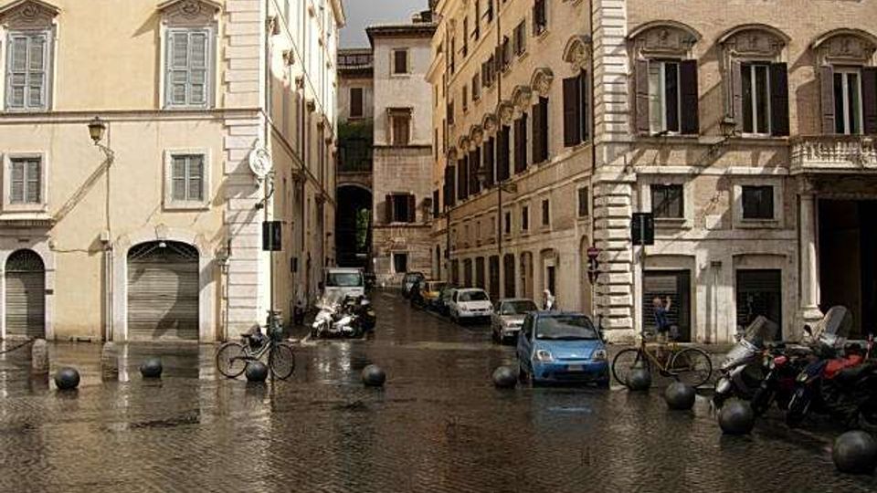Piazza urbana con pavimentazione in sampietrini bagnati, circondata da palazzi storici a più piani con facciate chiare, finestre con persiane e portici al piano terra. In primo piano ci sono dissuasori sferici, biciclette e veicoli parcheggiati lungo una strada stretta tra gli edifici.