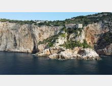 Scogliera costiera di roccia chiara a picco sul mare, con un promontorio roccioso coperto di macchia mediterranea. In alto sul crinale si vede una struttura in pietra simile a un forte o edificio storico, circondata da vegetazione.