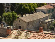 Vista dall’alto di un complesso di edifici in pietra con tetti in coppi e tegole, tra cui una struttura rettangolare con facciata semplice e piccola finestra circolare. Intorno si vedono alberi, una piazzetta lastricata e muretti con ringhiere ai margini.