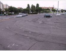 Ampio parcheggio asfaltato quasi vuoto con segnaletica a terra sbiadita e alcune auto lungo il bordo. Sullo sfondo si vedono alberi, una strada e edifici urbani in lontananza.
