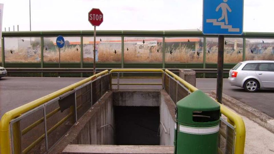 Ingresso a un sottopassaggio pedonale sotto una strada, con ringhiere gialle e scala che scende in un vano in cemento. Accanto ci sono un cestino verde e segnali stradali, con barriera antirumore trasparente sullo sfondo e auto sulla carreggiata.