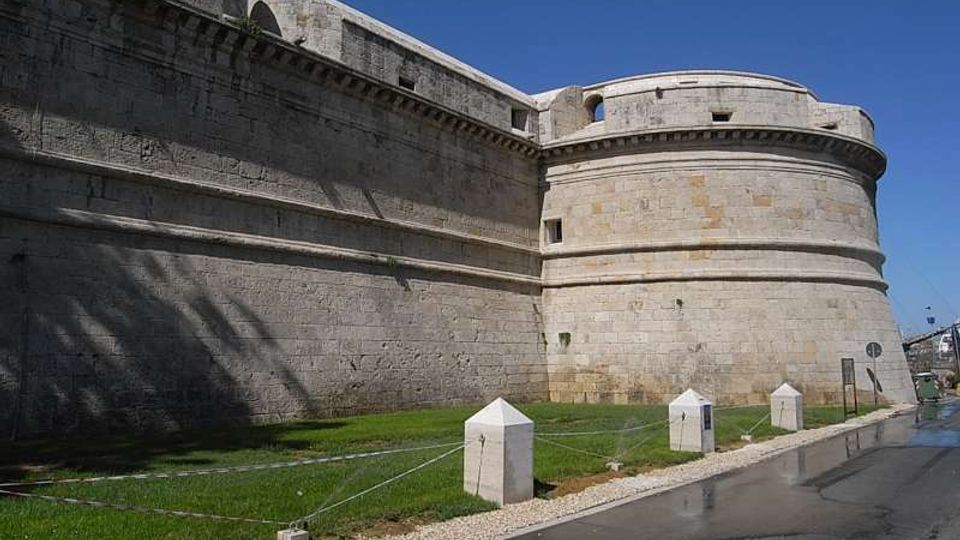 Mura esterne di una fortezza in pietra chiara con grande bastione cilindrico e cornici orizzontali. Davanti si vede una fascia di prato delimitata da paletti con cavi e una strada asfaltata lungo il perimetro sotto un cielo sereno.