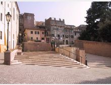 Scalinata in pietra che collega una piazzetta pavimentata in sampietrini a una terrazza con muretti e ringhiere. Sullo sfondo si vedono edifici storici con facciate usurate e finestre, lampioni in ferro e alberi al lato destro.
