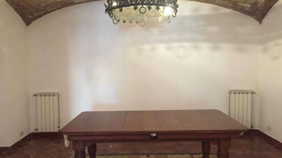 Interno di una sala con soffitto a volta in mattoni e pareti bianche, illuminata da un lampadario in ferro battuto. Al centro c’è un grande tavolo in legno sopra un tappeto, con pavimento in cotto e due radiatori ai lati.