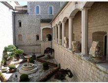 Chiostro in pietra con porticato ad archi su colonne lungo un lato, con capitelli e frammenti scultorei esposti sul parapetto. Nel cortile pavimentato si vedono aiuole bordate, vasi con piante e una fontana circolare centrale, circondati da mura in muratura con finestre ad arco.