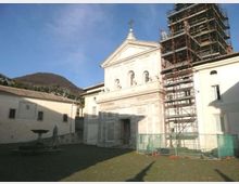 Facciata di una chiesa in una piazza con pavimentazione in pietra, con frontone triangolare e tre finestre ad arco; sul lato destro è presente un’alta impalcatura da cantiere con recinzione. Sullo sfondo si vedono edifici chiari e una collina sotto un cielo sereno.
