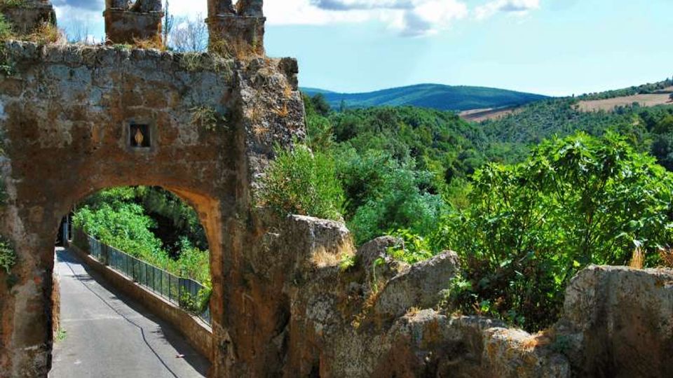 Antica porta in pietra con arco e merli, affacciata su una stretta strada asfaltata con ringhiera. Intorno si vedono rovine di mura e una valle verde con colline sullo sfondo sotto un cielo parzialmente nuvoloso.