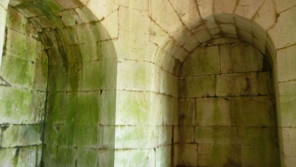 Interno di una struttura in pietra con pareti in blocchi e volte a botte, con due nicchie ad arco incassate e soffitto curvo. La superficie mostra macchie di umidità e patina verdastra, su pavimento in pietra.