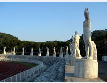 Anfiteatro all’aperto con gradinate in pietra che costeggiano una pista di atletica, affiancate da una fila di grandi statue bianche su piedistalli lungo il bordo superiore. Sullo sfondo si vedono colline alberate e cielo sereno.