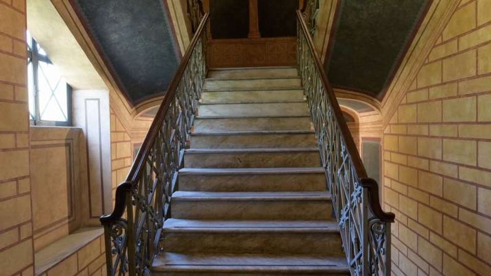 Scalinata interna in pietra che sale verso un pianerottolo, con ringhiere in ferro battuto su entrambi i lati. Pareti decorate con motivi geometrici, soffitto inclinato e pavimento a scacchiera in primo piano.