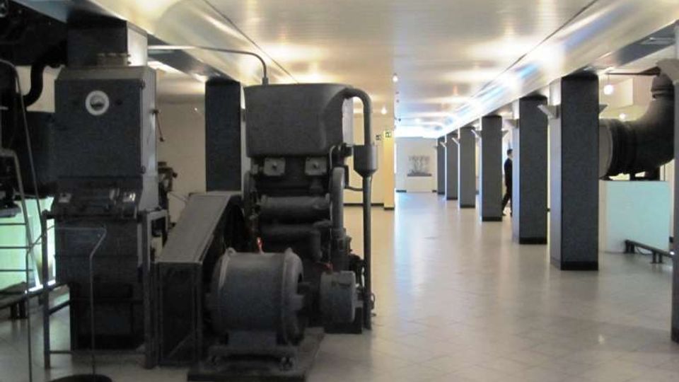 Ampio spazio espositivo interno con pavimento piastrellato e soffitto basso illuminato, scandito da pilastri scuri. Lungo il corridoio sono esposti grandi macchinari industriali e tubazioni metalliche.