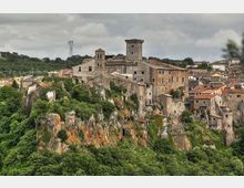 Borgo storico in pietra arroccato su una rupe, con case addossate e un grande edificio fortificato con torre quadrata e campanile. In primo piano si vedono le pareti rocciose e la vegetazione, con colline e cielo nuvoloso sullo sfondo.