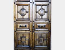 Portone in legno a doppia anta con pannelli intagliati geometrici e medaglioni circolari centrali, rifinito con borchie e maniglie in metallo scuro. Vista ravvicinata della superficie lignea con venature ben visibili e struttura simmetrica.