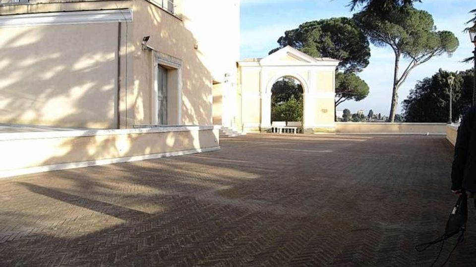 Cortile lastricato con pavimentazione in mattoni a spina di pesce, affiancato da un edificio chiaro con terrazza e finestre. Sullo sfondo si vede un arco monumentale e alberi alti in un’area esterna panoramica.