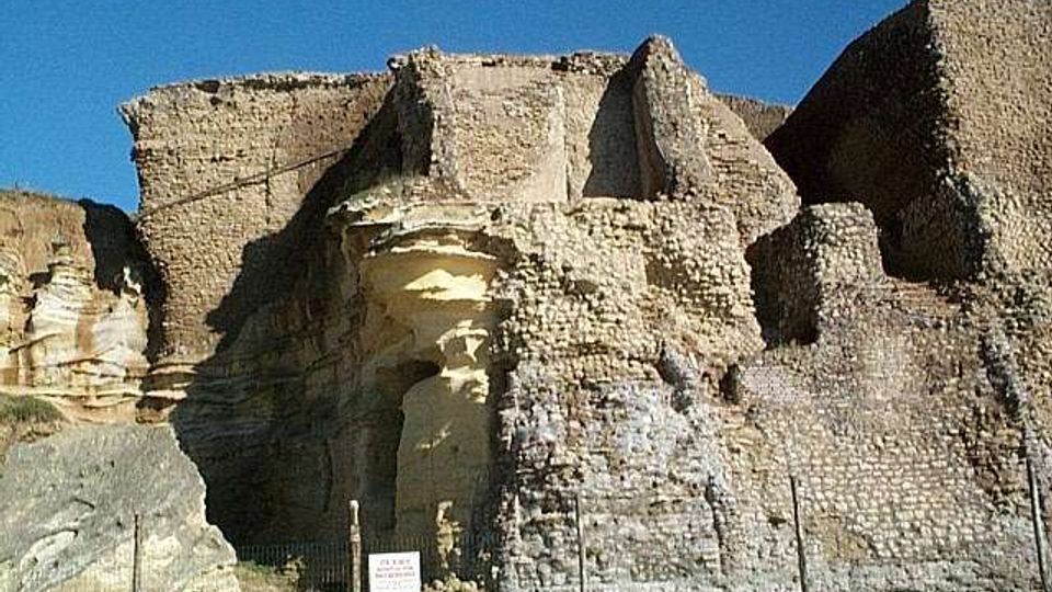Resti di una fortificazione in pietra con muri spessi e irregolari, addossata a una parete rocciosa chiara. Davanti si vede un’area arida recintata con pali e un piccolo cartello informativo sotto un cielo sereno.