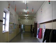 Corridoio interno di una scuola con pavimento piastrellato lucido, pareti bianche con fascia beige e disegni e cartelloni appesi. Dal soffitto pendono decorazioni colorate e lungo le pareti ci sono finestre, armadietti e appendiabiti con giacche; in fondo si vede una porta aperta.