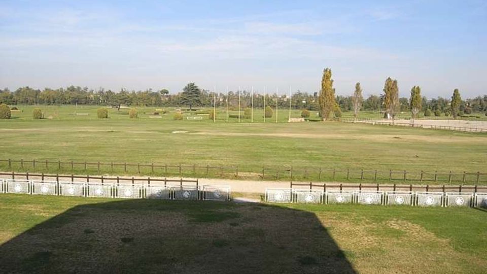 Ampia area verde pianeggiante con prati e recinzioni, con un sentiero sterrato in primo piano e file di alberi e pali al centro. Sullo sfondo si vede un paesaggio aperto con vegetazione sparsa sotto un cielo sereno.