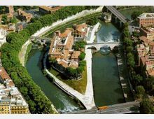 Veduta aerea di un’isola urbana triangolare circondata da un fiume, con edifici storici dai tetti in terracotta e argini in pietra. Due ponti attraversano il corso d’acqua e una piccola cascata è visibile vicino a uno degli sbarramenti, con alberi e strade che costeggiano le rive.