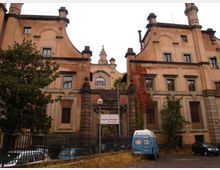 Facciata di un grande edificio storico in mattoni, simmetrico, con due corpi laterali e una parte centrale arretrata con frontone e finestrone ad arco. Davanti si vede un cancello in ferro, vegetazione rampicante e un’area di accesso con alcuni veicoli parcheggiati.