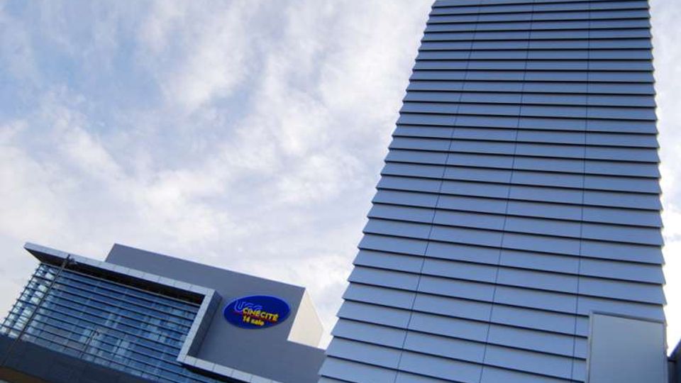 Ripresa dal basso di un edificio moderno con facciata in pannelli orizzontali chiari, affiancato da una struttura vetrata. Sul lato sinistro si vede un’insegna circolare su una parete grigia, con cielo parzialmente nuvoloso sullo sfondo.