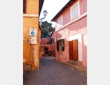 Vicolo pedonale acciottolato tra edifici a due piani intonacati in tonalità rosa e arancione, con persiane e portone in legno. Sulla parete sinistra sono visibili un lampione a muro e un cartello “area pedonale”, mentre il vicolo conduce a un piccolo cortile con ingresso ad arco.