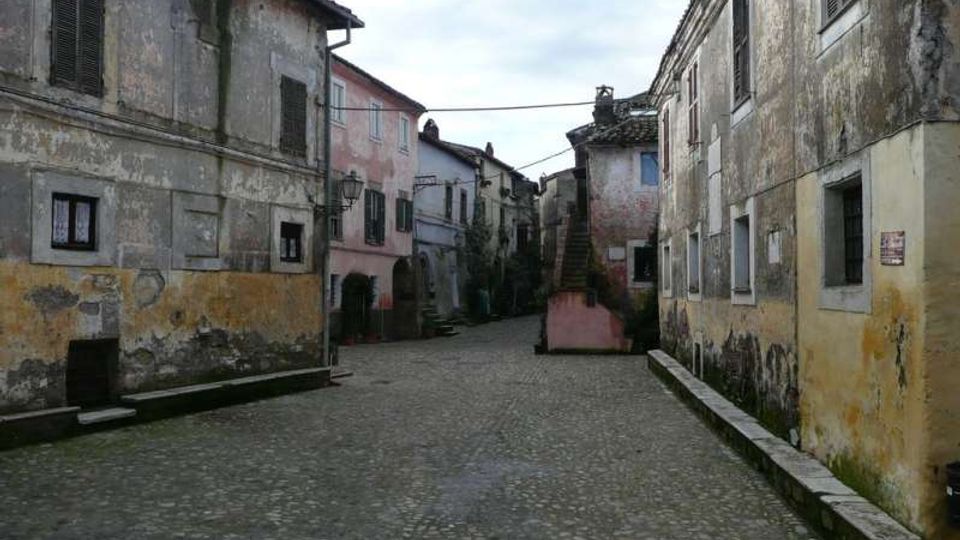 Strada in acciottolato in un borgo storico, fiancheggiata da case in muratura intonacata e consumata dal tempo con persiane scure. In fondo si vedono un arco di passaggio, una piccola scalinata esterna e cavi sospesi tra gli edifici.
