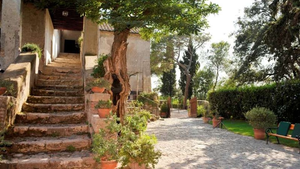 Cortile esterno in pietra con scala di accesso a un edificio rustico intonacato, affiancata da vasi in terracotta con piante. Viale pavimentato che prosegue tra alberi e siepi, con panchina e ingresso con cancello sullo sfondo.