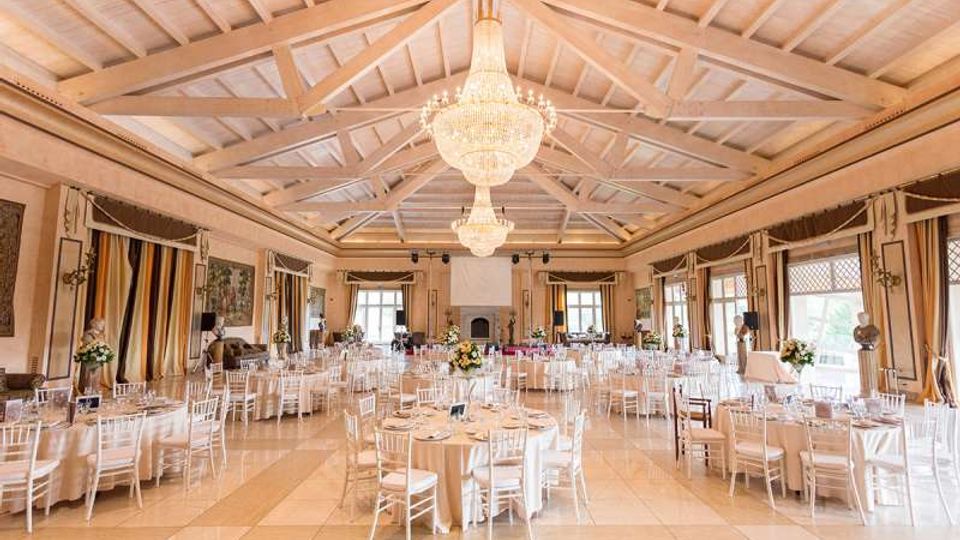 Ampia sala per eventi con soffitto alto a capriate in legno bianco e grandi lampadari a goccia centrali. Tavoli rotondi apparecchiati con sedie bianche, ampie finestre con tende laterali e camino sul fondo della sala.