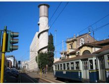 Strada urbana con binari e linee elettriche del tram, con un tram blu in primo piano. Sullo sfondo si vede un alto edificio industriale con grande ciminiera cilindrica e rampa elicoidale esterna, accanto a edifici storici con tetti in coppi e facciate in muratura.