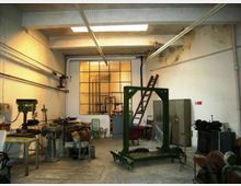 Interno di un’officina industriale con pavimento in cemento, macchinari e banchi da lavoro, tubazioni a vista e lucernario sul soffitto. Sul fondo una grande finestra a griglia e una scala appoggiata vicino ad armadi metallici e attrezzature.