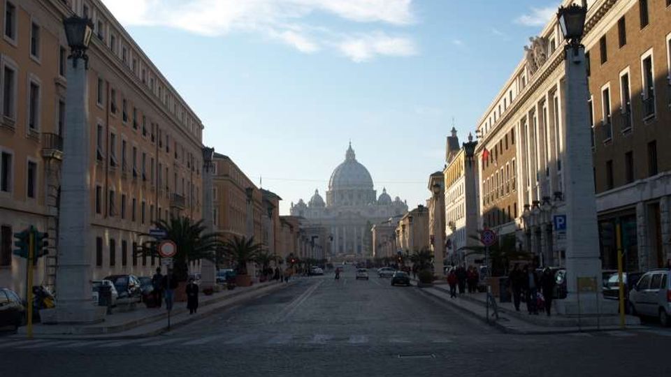 Lungo viale urbano rettilineo con carreggiata ampia, fiancheggiato da edifici storici e lampioni monumentali, che conduce in prospettiva verso la cupola della Basilica di San Pietro sullo sfondo. Sul bordo della strada si notano marciapiedi, alcune palme e auto parcheggiate.
