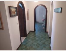 Corridoio interno di una casa con pareti bianche e pavimento in piastrelle verdi decorate, che conduce a due porte ad arco con cornici in legno scuro. Alle pareti sono appesi quadri e in fondo si intravedono un tavolino bianco con lampada e un armadio.