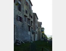 Fila di edifici storici in pietra con persiane scure e piccole finestre, affacciati su un prato lungo una stretta riva murata. Sul lato destro si intravede un parapetto in pietra e un tratto di panorama in lontananza.