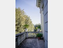 Terrazza esterna di un edificio classico con pavimento piastrellato e balaustra in pietra, affacciata su alberi alti. Vasi con piante ornamentali sono disposti lungo il parapetto e vicino alla parete con cornici decorative.