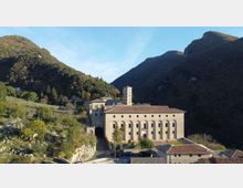 Complesso monastico in pietra con tetti in tegole e una torre campanaria quadrata, situato ai piedi di montagne boscose. In primo piano si vede un cortile con edifici annessi e una strada d’accesso, circondati da vegetazione e pendii rocciosi.