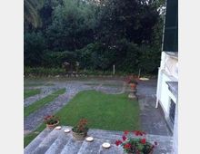 Cortile esterno con prato curato e vialetti in pietra, circondato da alberi e siepi. In primo piano ci sono gradini con vasi di fiori e, sul lato destro, la facciata di una casa con persiane verdi.