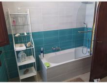 Bagno con vasca bianca dotata di parete in vetro e soffione doccia, rivestito da piastrelle bianche e una fascia turchese a effetto ondulato. Accanto alla vasca c’è una scaffalatura bianca con ripiani in vetro e accessori decorativi, e una porta in legno sul lato destro.