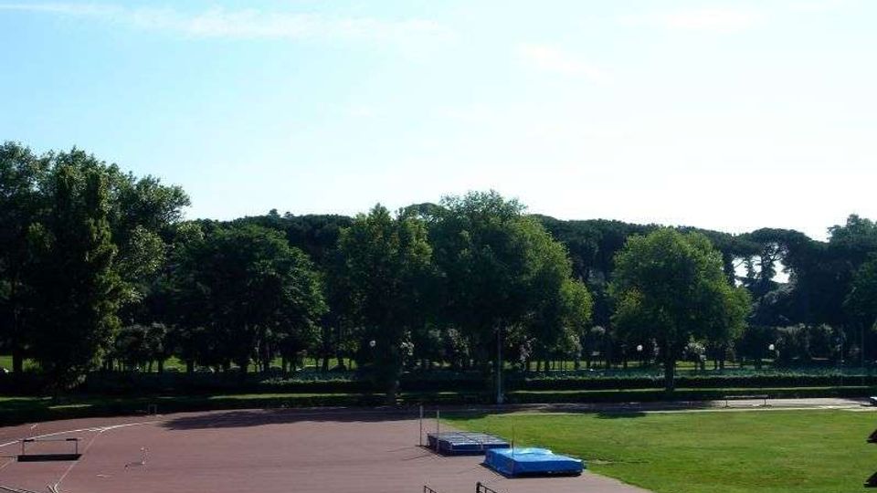 Campo sportivo all’aperto con pista di atletica in tartan che circonda un’area in erba. Sul lato si vede una pedana con materassi per salti e, sullo sfondo, una fila di alberi sotto un cielo sereno.