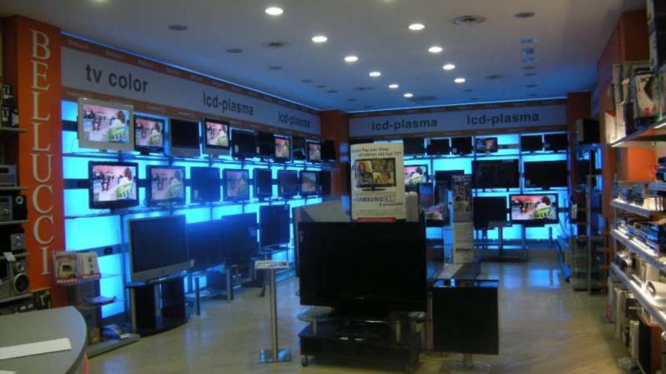 Interno di un negozio di elettronica con pareti attrezzate da numerosi televisori e monitor esposti, illuminati da pannelli retroilluminati azzurri. Soffitto con faretti incassati, pavimento chiaro e scaffalature laterali con prodotti e accessori.